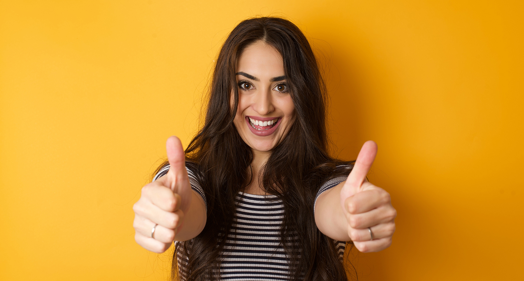 Portrait of a happy smiling blue eyed young successful woman giving two thumbs up gesture standing indoors. Positive human emotion facial expression body language.